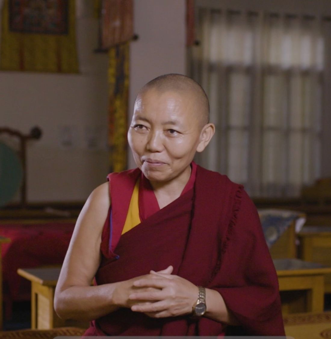A Geshe-ma in maroon robes, hands clasped, seated indoors, one of the first women to earn Tibetan Buddhism's highest degree, supported by The Maitri Project