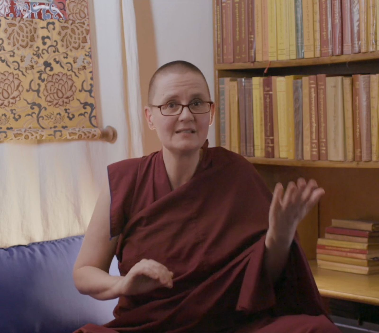 A Buddhist nun in maroon robes speaking, framed by a wall of scripture books